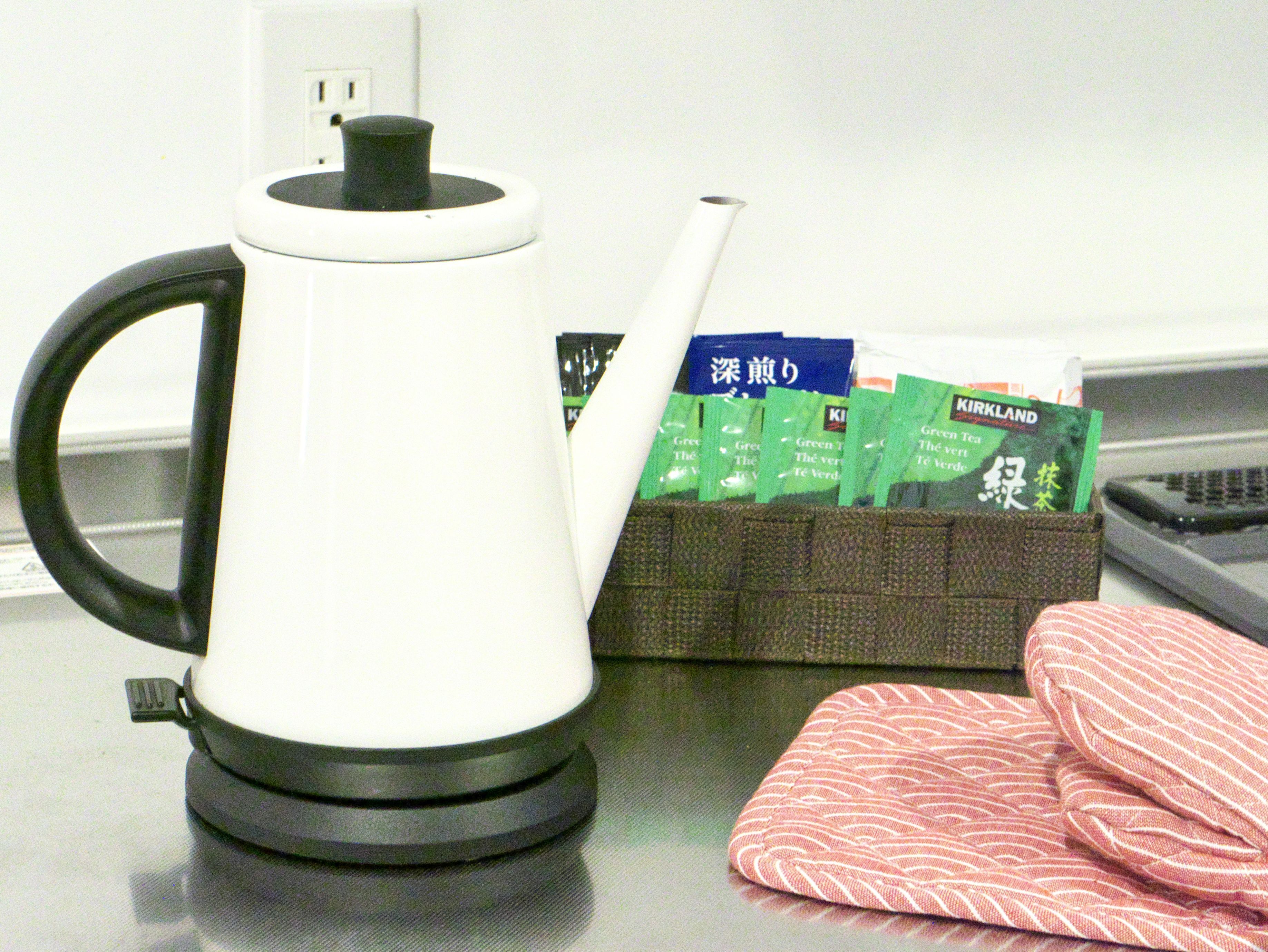 Una tetera eléctrica blanca sobre una mesa, junto a una cesta marrón que contiene bolsitas de té verde y un objeto blanco.