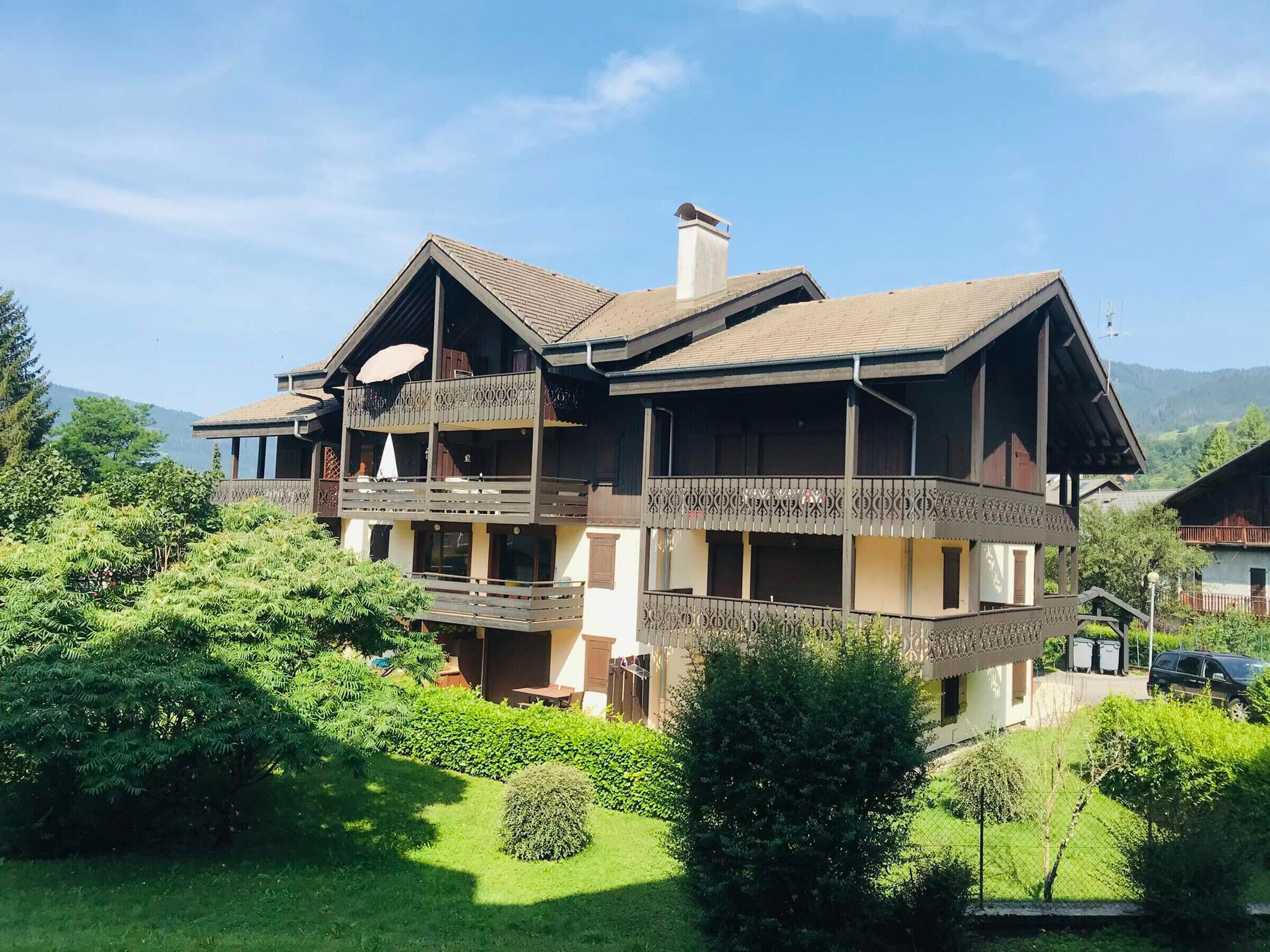 A two-story chalet with a balcony, surrounded by lush greenery and mountains in the background.