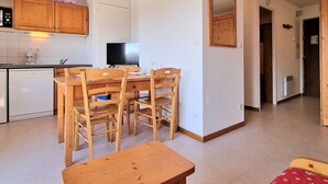 A compact kitchen with wooden cabinets, a dining table with chairs, a television, and a small refrigerator.