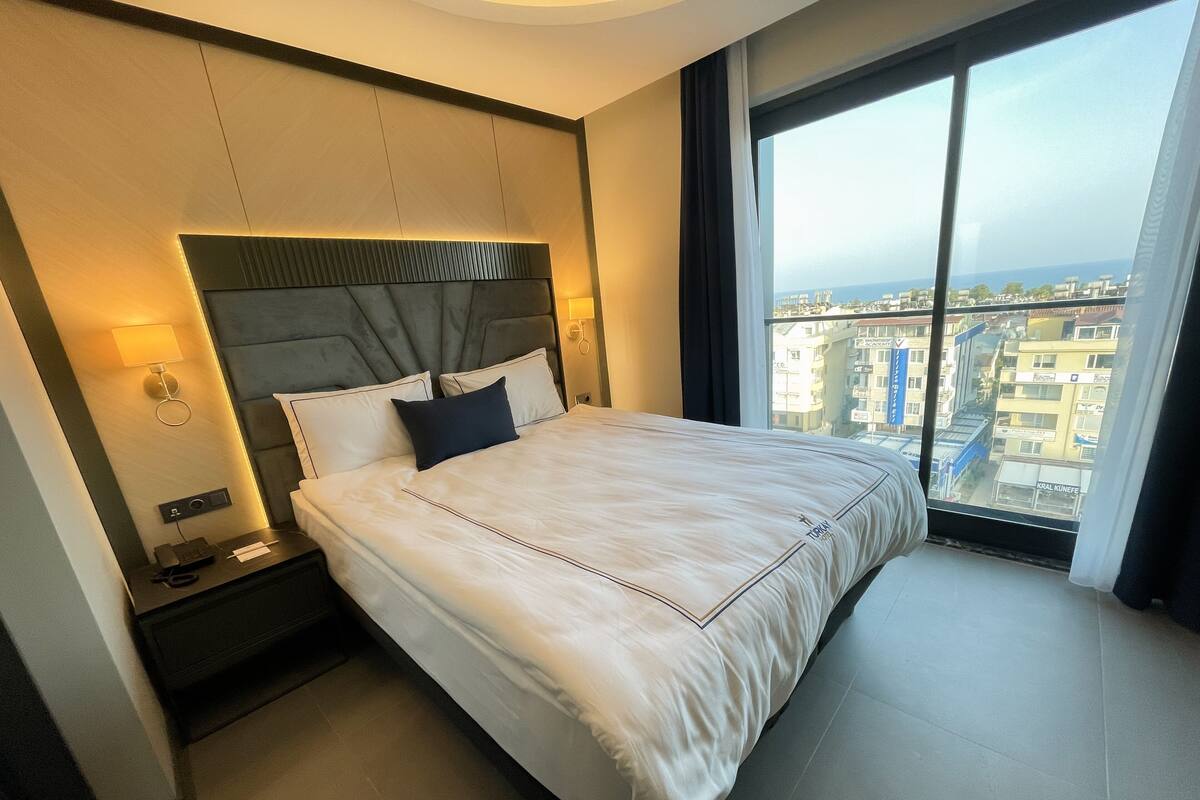 Ein Hotelzimmer mit einem großen Bett, einem Nachttisch mit Lampe und Blick auf die Stadt durch ein großes Fenster.