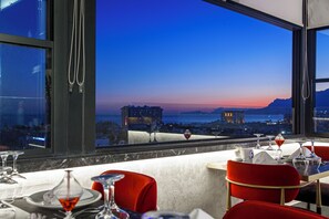 A restaurant with a table set for two, a view of the cityscape, and a sunset over the water. - Turkay Hotel (Konyaalti)