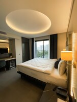 A modern hotel room with a large bed, a desk, a minibar, and a view of the city.