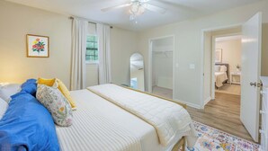 Une chambre à coucher comprenant un lit, un ventilateur de plafond, une fenêtre avec des rideaux, une porte donnant sur une autre pièce et un petit tapis.