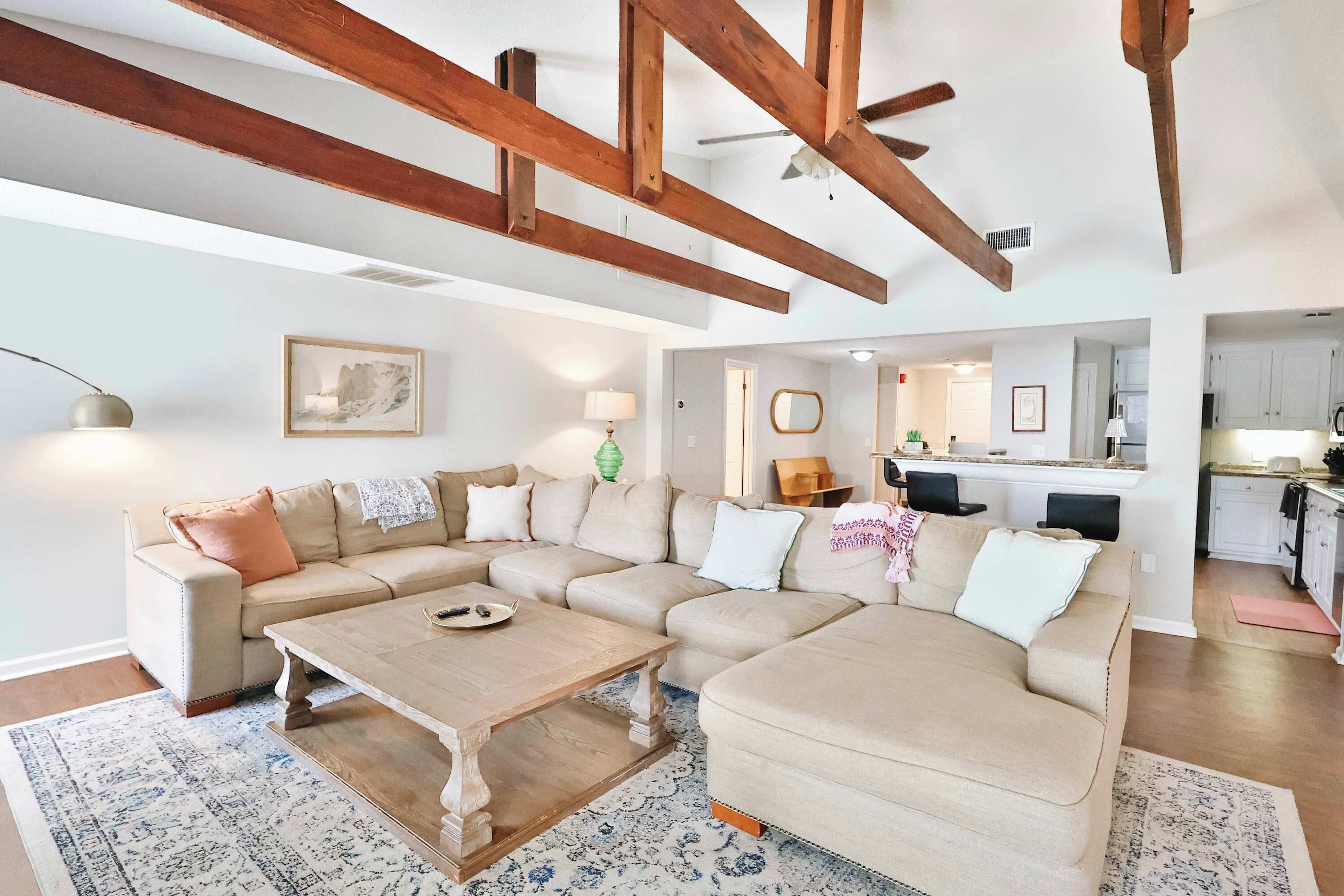 A living room with a large L-shaped beige sofa, a wooden coffee table, and a patterned rug. There is a kitchen visible in the background.