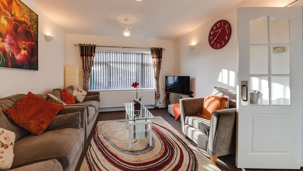 A living room with a sofa, armchair, and a coffee table. There is a large window with curtains, a wall clock, and a television. - 3 Bedroom Home. Home Working. Free Parking. Garden. Wigan (Wigan)