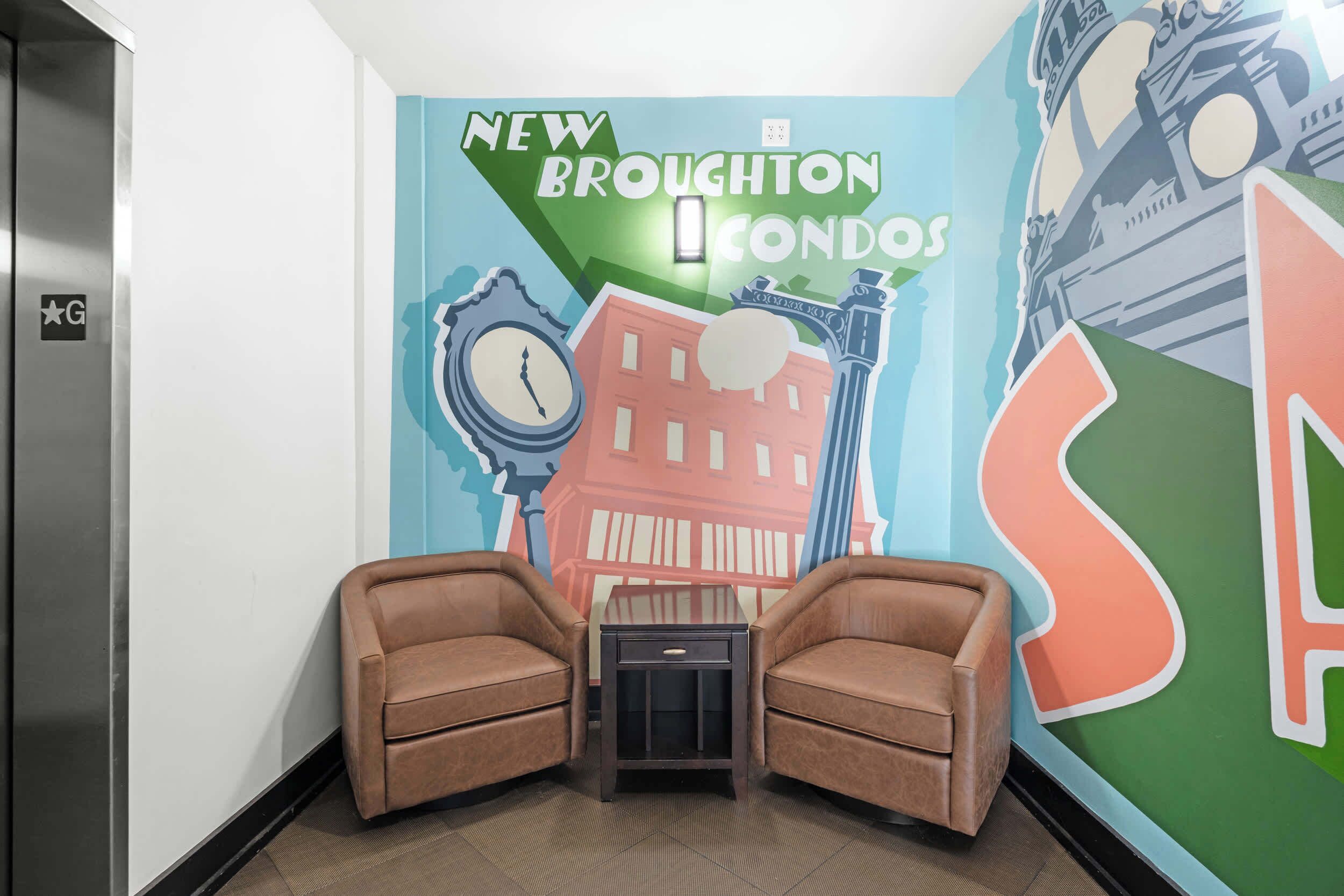 A lobby with two brown leather armchairs, a small table, and a wall with colorful artwork depicting a clock, a building, and the text "NEW BROUGHTON ICONOS".