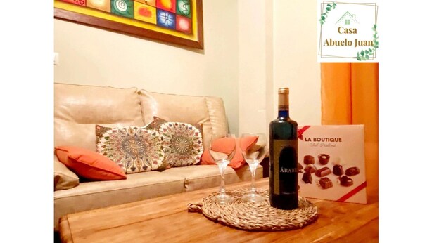 Un salon avec un canapé, des coussins décoratifs, une bouteille de vin, des verres à vin et un set de table sur une table en bois.