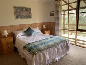 A bed with a white comforter and a blue and green woven blanket, wooden headboard, two bedside lamps, a wooden nightstand with a drawer, a framed painting above the bed, and a window with a view of trees. - Mansfield Colonial Cabin with Stunning Views (Mansfield)