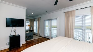 A bedroom with a bed, a ceiling fan, a TV, and a view of the outdoors through large windows. - Steel Waters – Luxury Deep-Water Retreat with Private Dock & Stunning Marsh Views (Edisto Beach)
