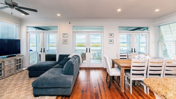A spacious living room with a large gray sectional sofa, a wooden dining table with white chairs, and a television on a wooden cabinet. - Steel Waters – Luxury Deep-Water Retreat with Private Dock & Stunning Marsh Views (Edisto Beach)