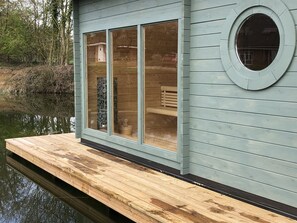 Une jetée en bois s’étendant dans une étendue d’eau calme, avec un bâtiment doté de grandes fenêtres et d’une fenêtre circulaire sur le côté droit.