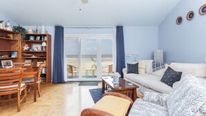A living room with a sofa, chairs, a dining table, and a bookshelf. There is a large window with curtains, and a ceiling fan. - Quail Hollow by Distinctive Beach Rentals (St. Augustine)