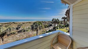 A balcony with a wicker chair, overlooking a beach with palm trees and a clear blue sky. - Quail Hollow by Distinctive Beach Rentals (St. Augustine)