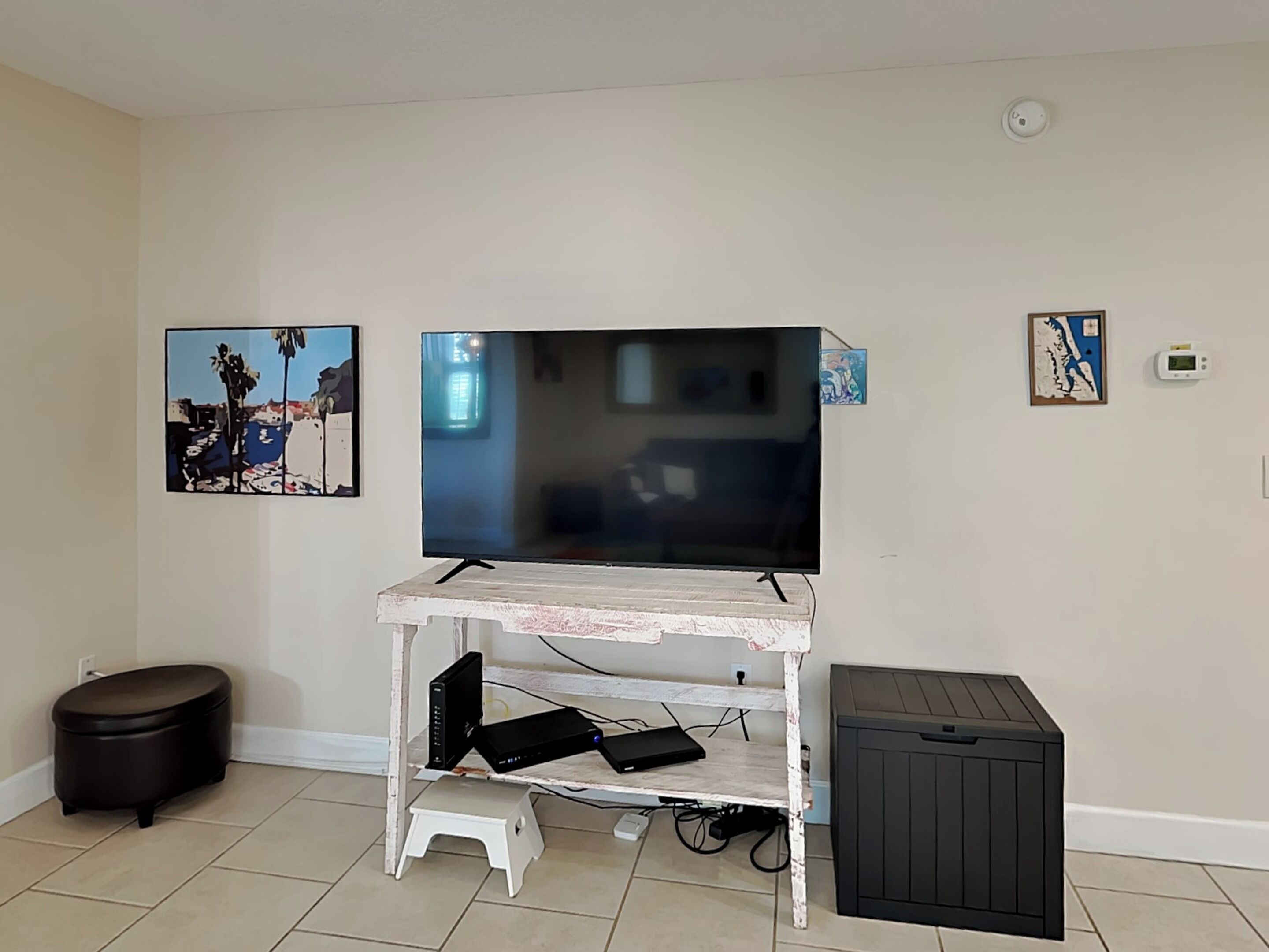 Un salon avec un téléviseur posé sur un meuble blanc, un petit tabouret, une poubelle et deux tableaux encadrés accrochés au mur.