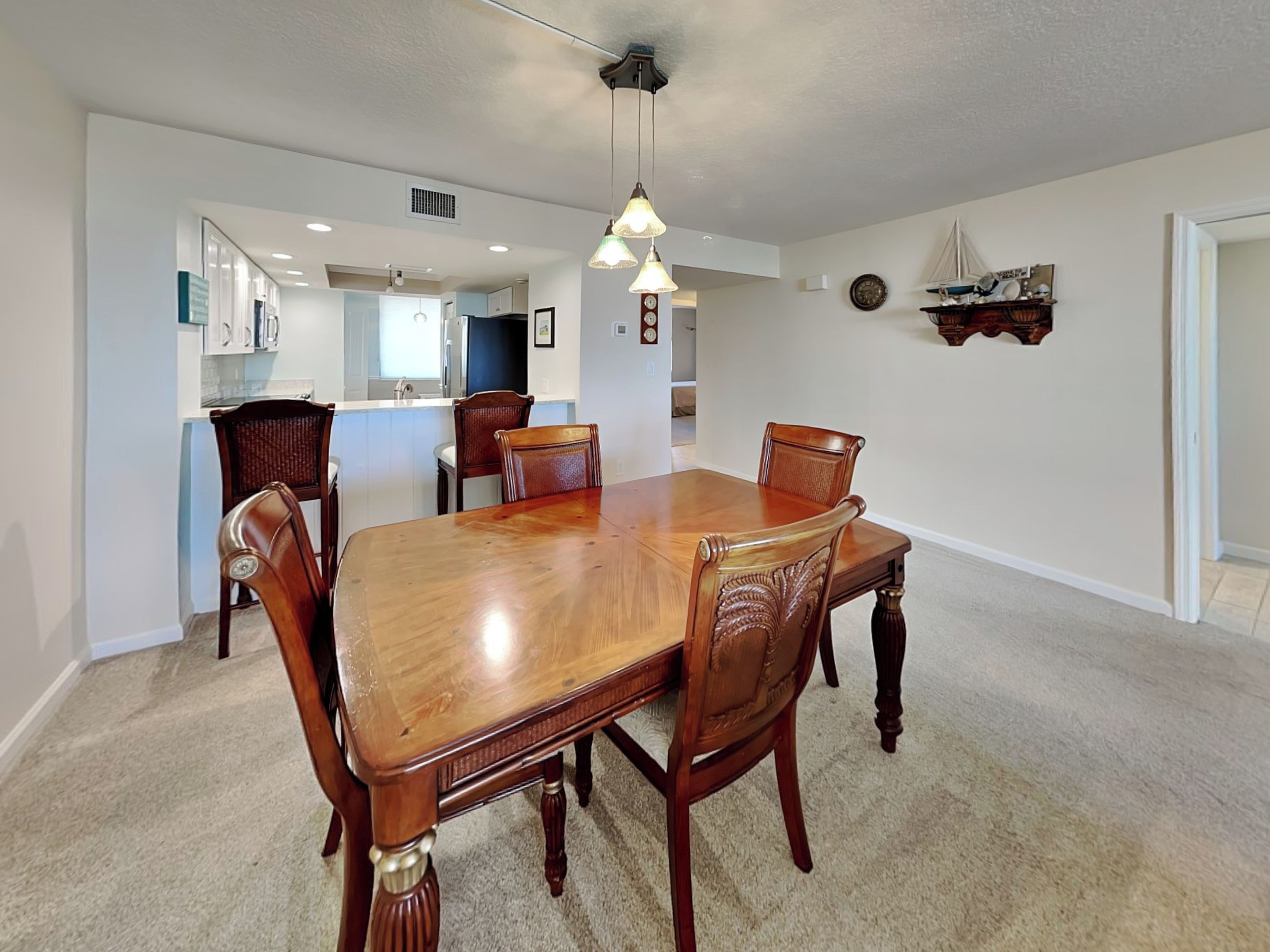 A dining area with a wooden table and chairs, a kitchen in the background, and a clock on the wall.