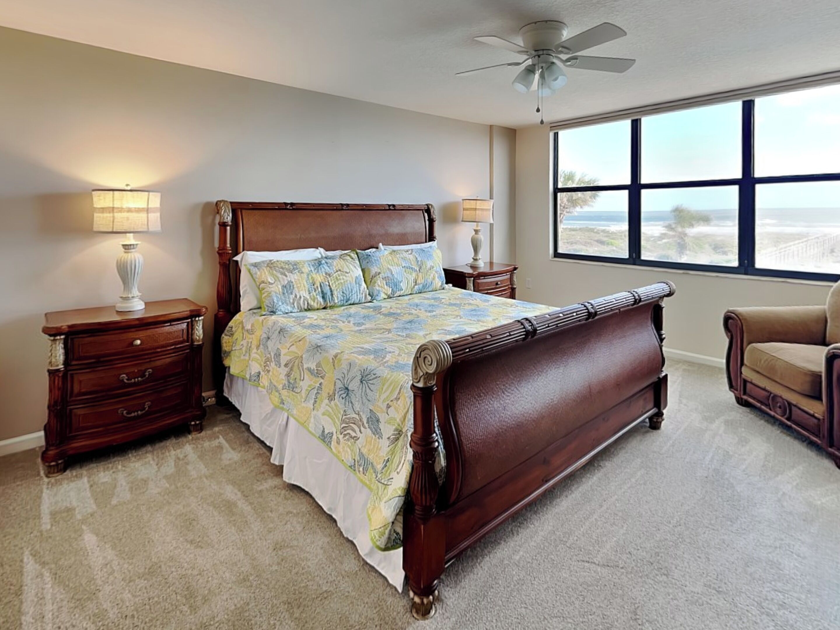 A bedroom with a large bed, two bedside tables, a chair, a ceiling fan, and a large window with a view.