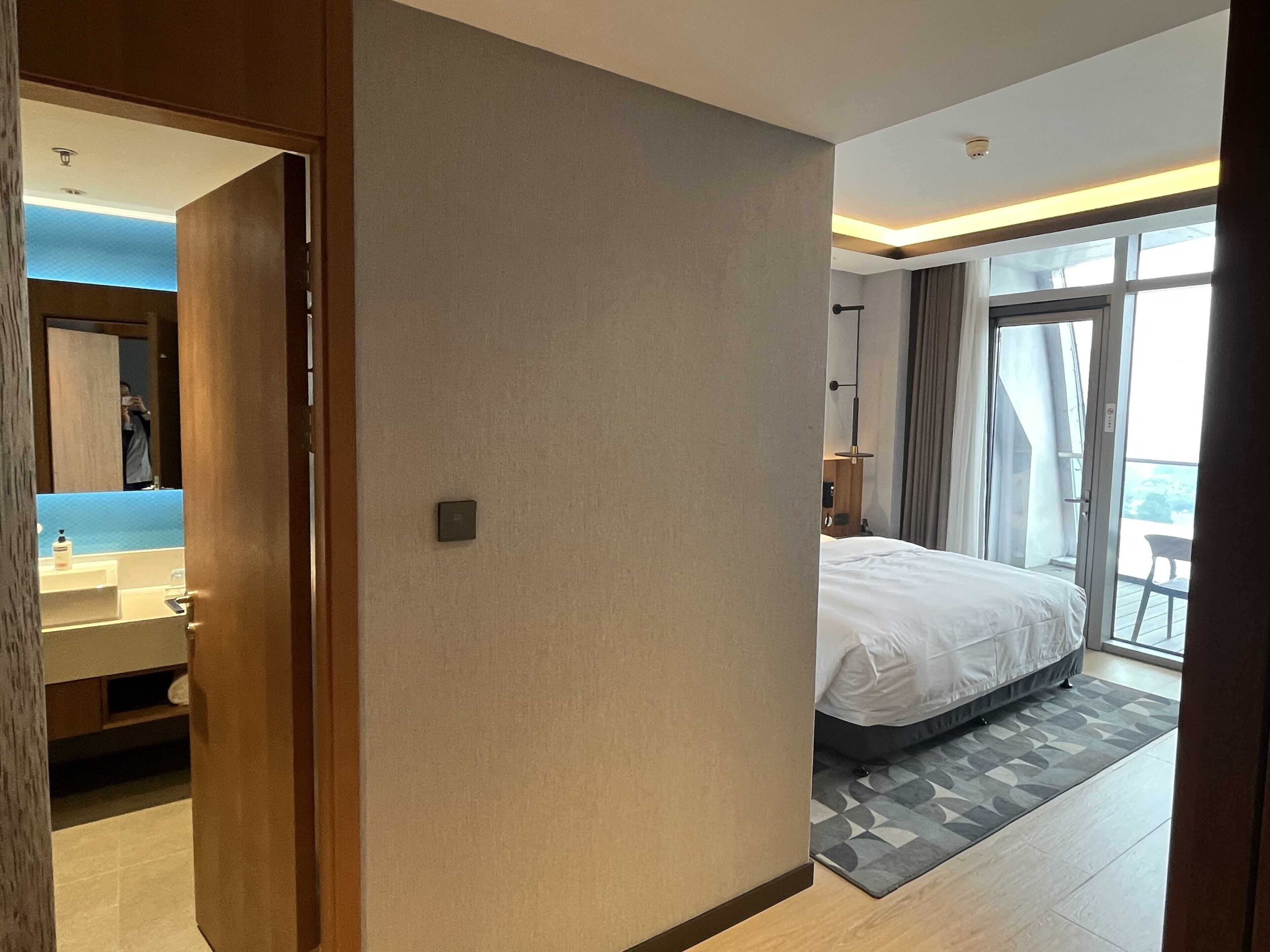 A hotel room with a bed, a bathroom with a sink and mirror, and a balcony with a view.