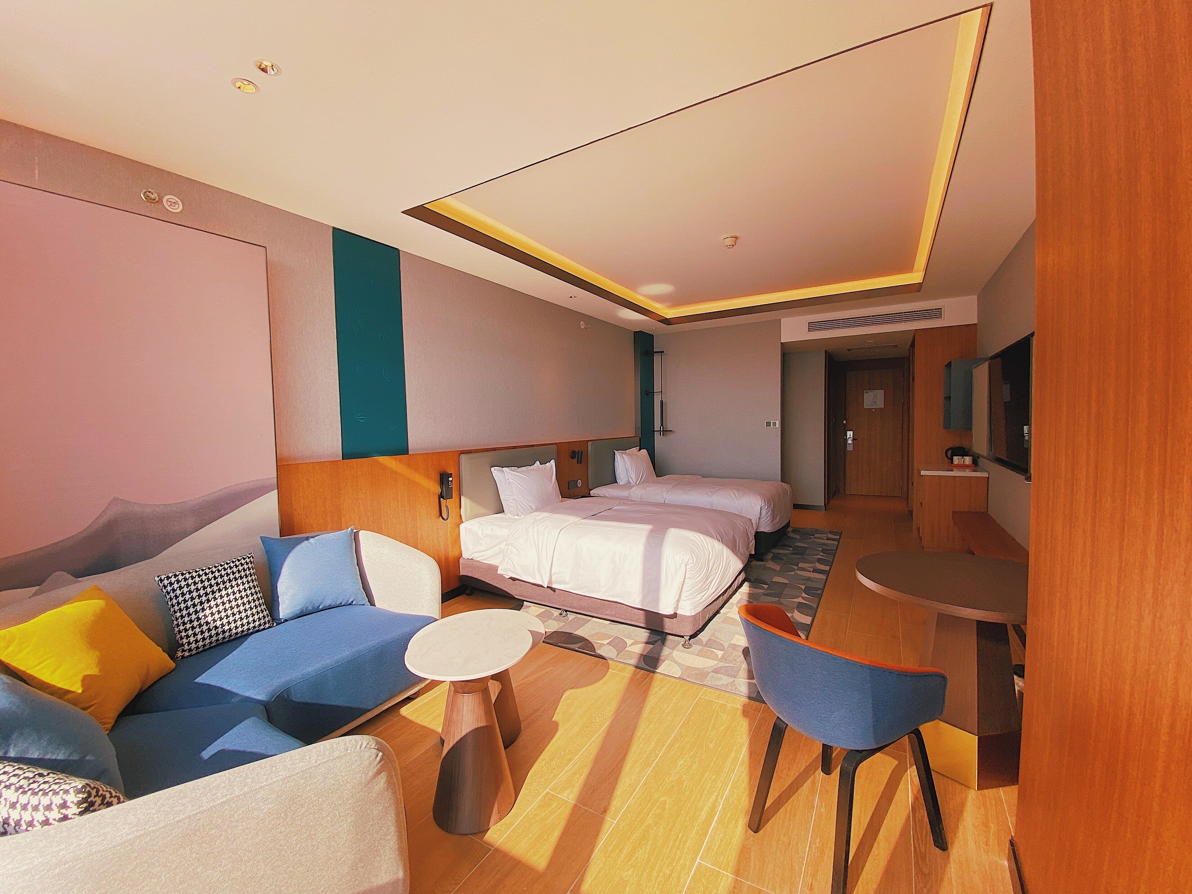 A modern hotel room with a sofa, two beds, a dining area, and a bathroom.