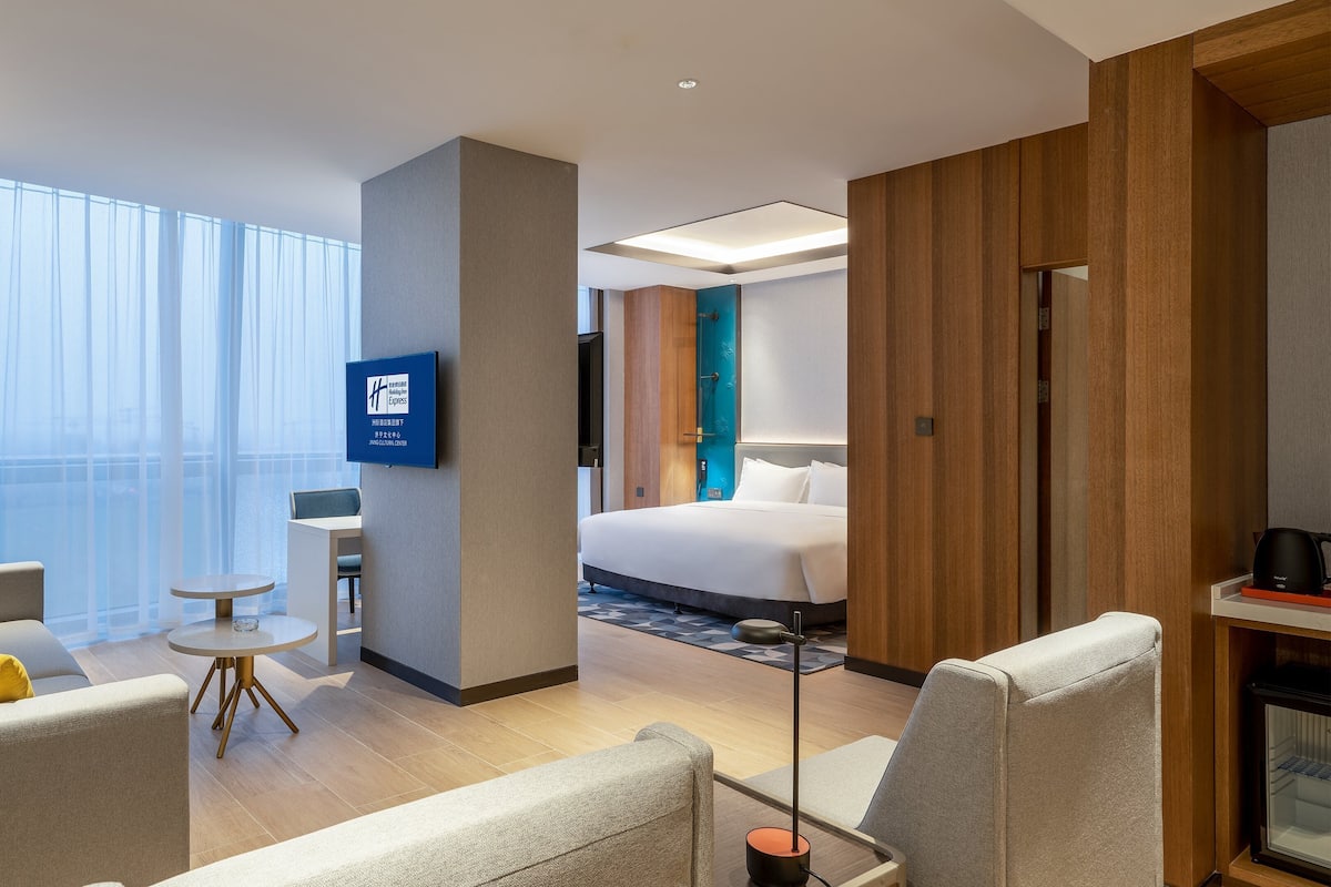 A modern hotel room with a large bed, a sitting area with a small table and chairs, a TV mounted on the wall, and a view of the city through large windows.
