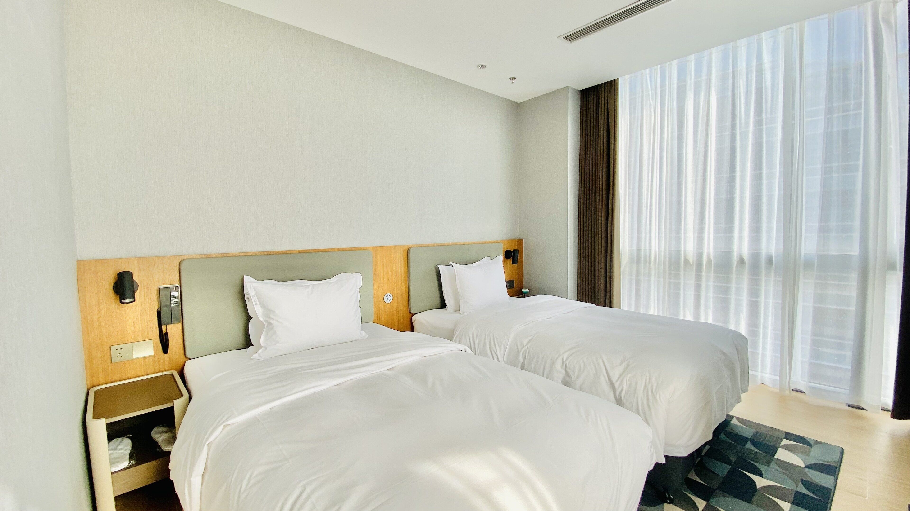 Quarto de hotel com duas camas, cabeceira de madeira, mesa de cabeceira com telefone e tomada, e uma janela grande com cortinas finas.