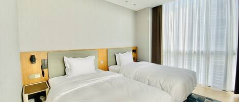Quarto de hotel com duas camas, cabeceira de madeira, mesa de cabeceira com telefone e tomada, e uma janela grande com cortinas finas.