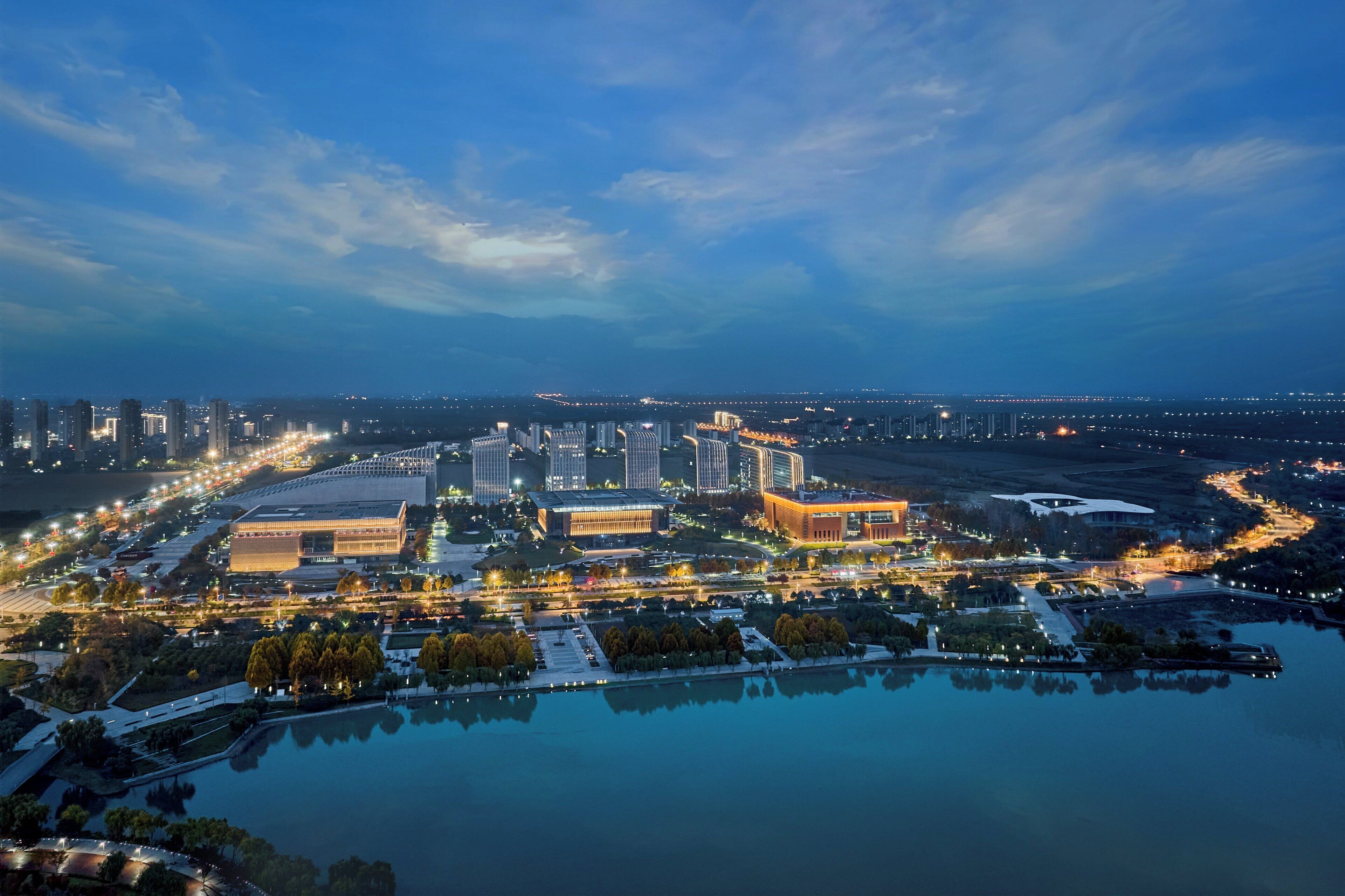 An aerial view of a cityscape with modern buildings, a large body of water, and a well-lit urban area.