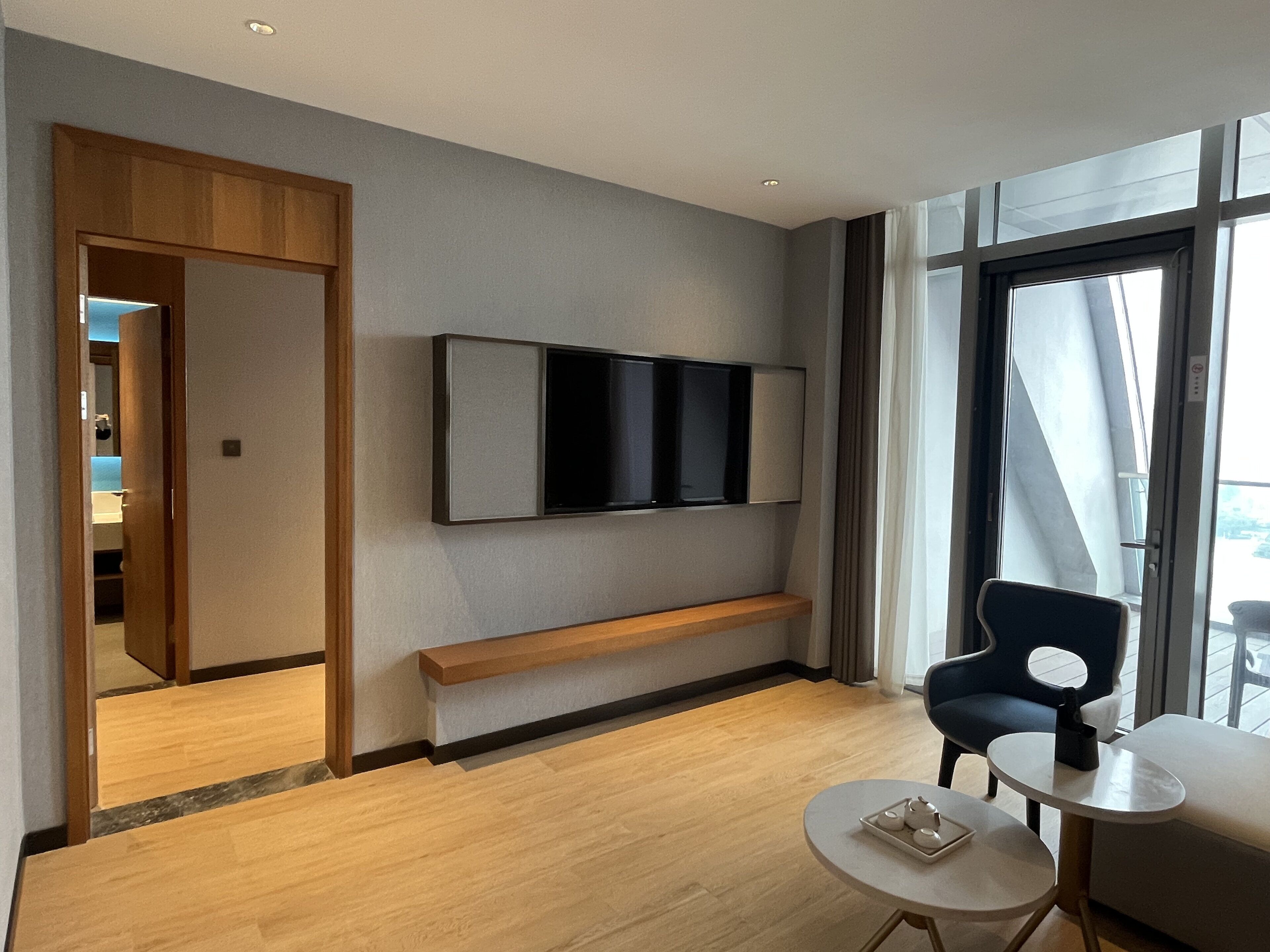 Una habitación de hotel moderna con un televisor grande montado en la pared, un banco de madera, una mesa redonda con una bandeja de té y vistas al exterior a través de amplios ventanales.