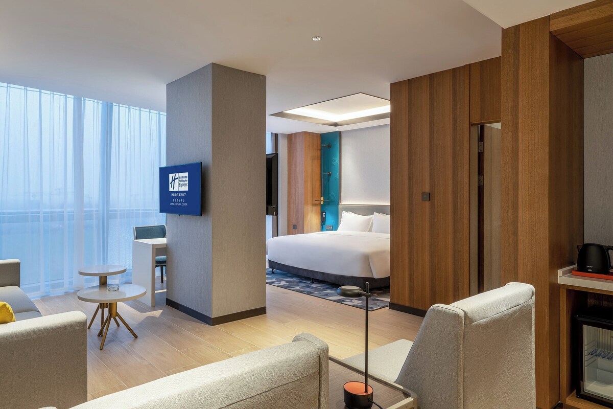 A hotel room with a bed, a desk, a chair, a small table, and a television.