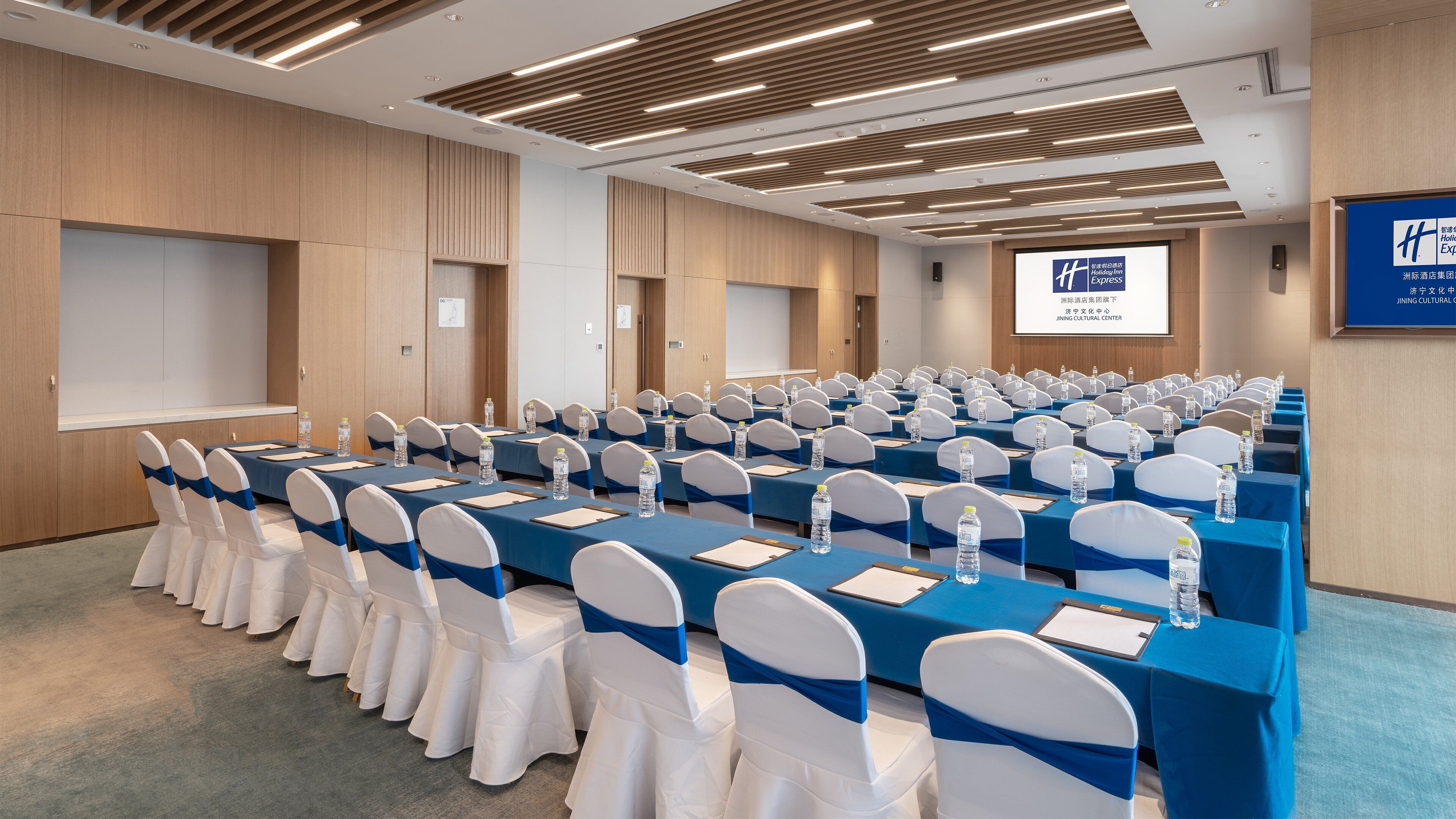 A conference room with rows of tables and chairs, equipped with water bottles, and a projection screen on the wall.