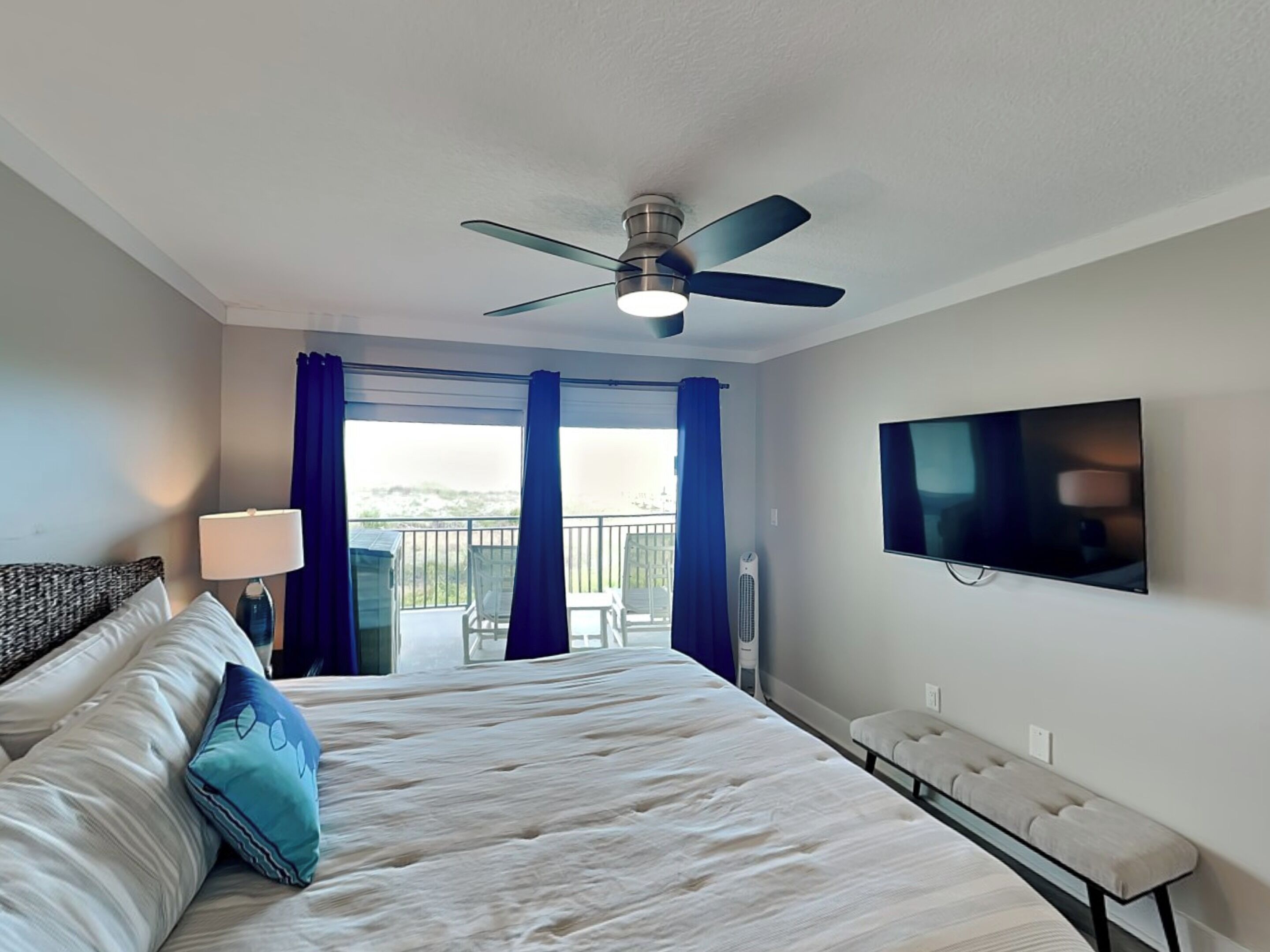 A hotel room with a bed, a ceiling fan, a TV, and a balcony with curtains.