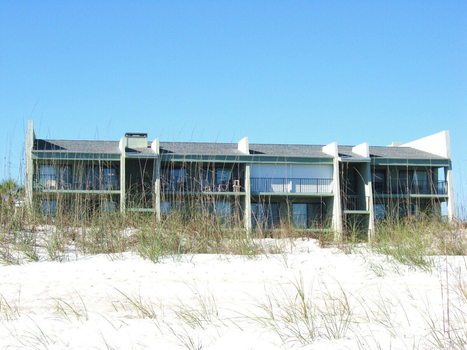 A multi-story building with a flat roof, large windows, and balconies. It is surrounded by sand and sparse vegetation.