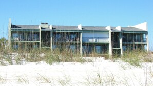 Ein mehrstöckiges Gebäude mit flachem Dach, großen Fenstern und Balkonen. Es ist von Sand und spärlicher Vegetation umgeben.