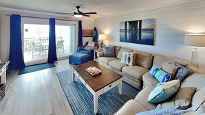 Ein Wohnzimmer mit einer Couch, einem Couchtisch und einem Esstisch. Es gibt einen Deckenventilator, ein Bild an der Wand und einen Blick ins Freie durch eine Schiebetür.
