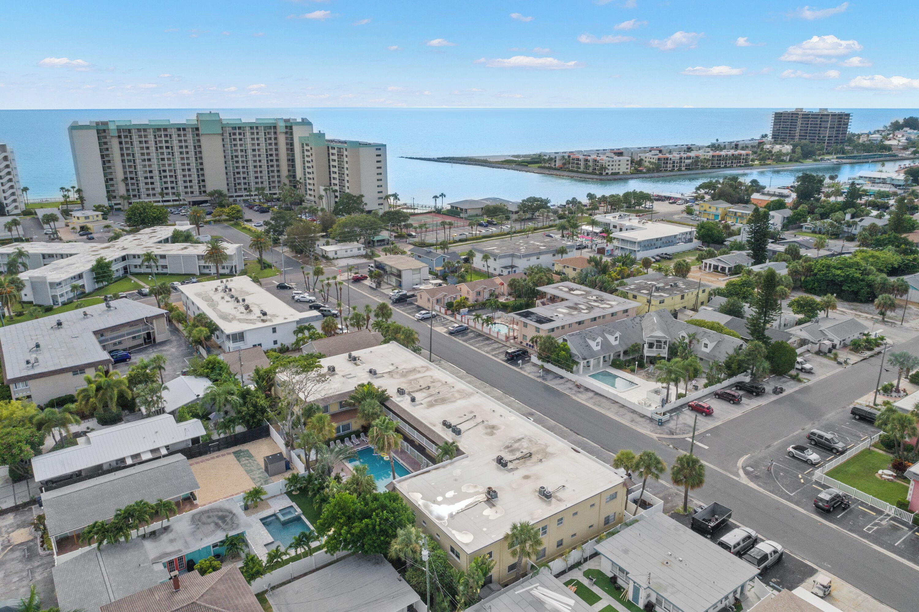 An aerial view of a coastal urban area with a mix of residential and commercial buildings, a prominent hotel with a large pool, and a marina in the background.