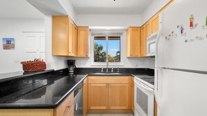 A modern kitchen with wooden cabinets, a black countertop, a stainless steel refrigerator, and a window with a view of palm trees. - Waves by Distinctive Beach Rentals (St. Pete Beach)