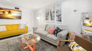 A modern living room with a yellow sofa, grey sofa, and a coffee table. There are three framed pictures on the wall and a hanging light fixture. - Waves by Distinctive Beach Rentals (St. Pete Beach)