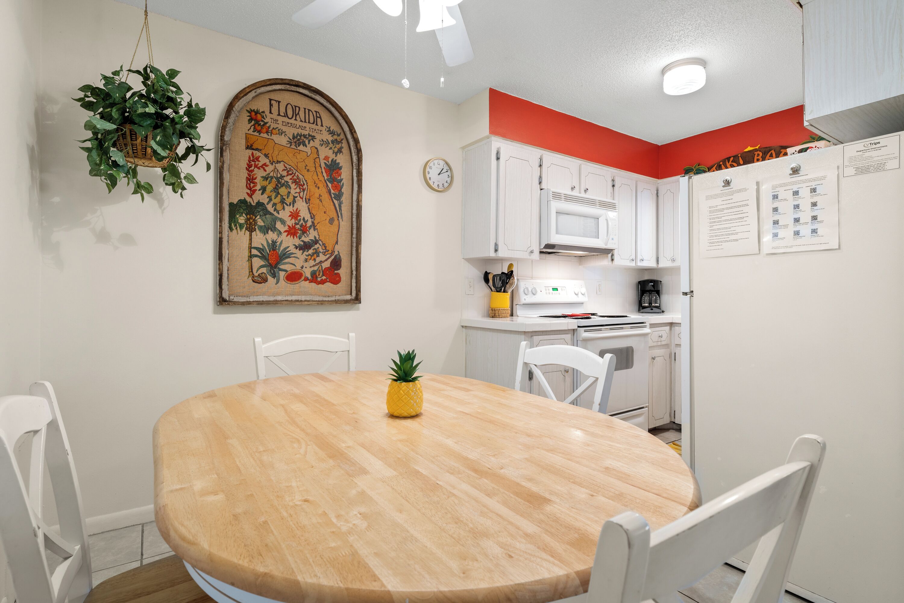 A dining area with a wooden table, white chairs, a pineapple centerpiece, a hanging plant, and a vintage floral poster on the wall.