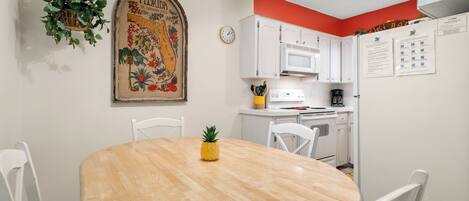 A dining area with a wooden table, white chairs, a pineapple centerpiece, a hanging plant, and a vintage floral poster on the wall.