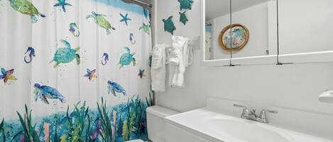 Un baño con cortina de ducha con tortuga marina, un inodoro blanco, un lavamanos con espejo y un perchero para toallas.