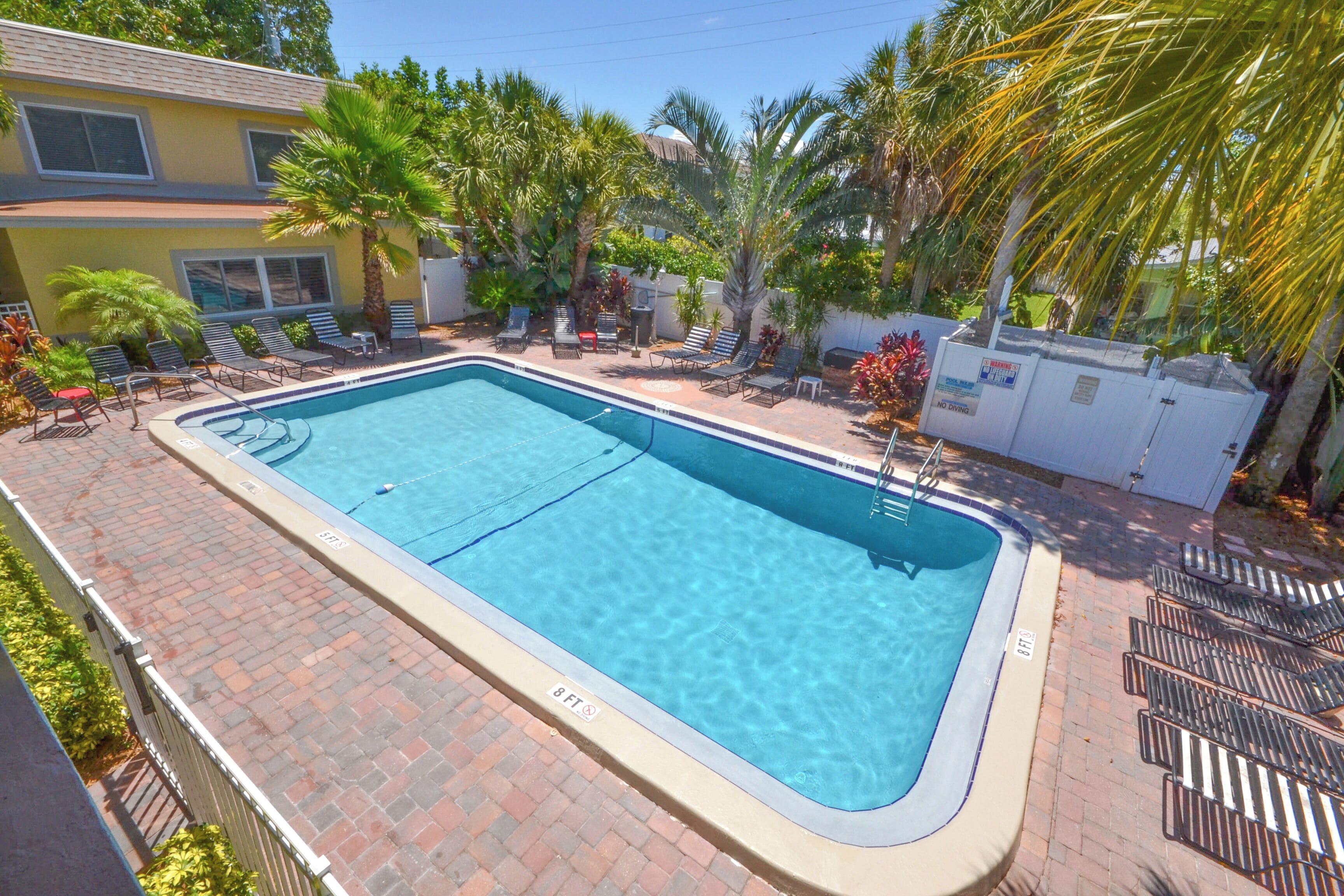 Une piscine rectangulaire bordée d’une terrasse en briques, entourée de chaises longues et de palmiers.