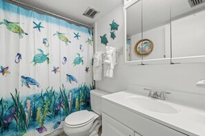 A bathroom with a sea turtle shower curtain, a white toilet, a sink with a mirror above it, and a towel rack with white towels. - Waves by Distinctive Beach Rentals (St. Pete Beach)
