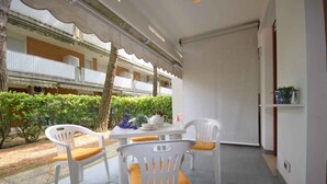 Un espacio de estar al aire libre cubierto con sillas de plástico blanco y una mesa, rodeado de setos y un edificio con balcones al fondo.