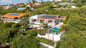 Une maison moderne dotée d’une piscine, entourée d’une végétation luxuriante et surmontée d’un toit équipé de panneaux solaires.