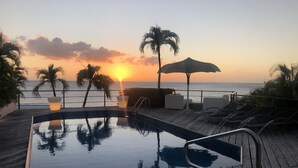 Une terrasse en bois avec une piscine, des chaises longues et une vue sur le coucher de soleil sur l’océan.