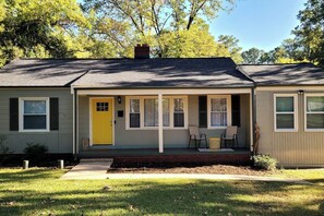 A single-story house with a yellow front door, a covered porch with two chairs, and a brick chimney. - 4BR Getaway | King Bed | Prime Uptown-SouthEnd (Charlotte)