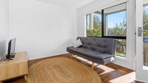 A modern living room with a grey sofa, a wooden coffee table, and a large window offering a view of greenery outside. - OSPREY - MORNINGTON. Walking distance to Mornington's cafe culture. (Mornington)