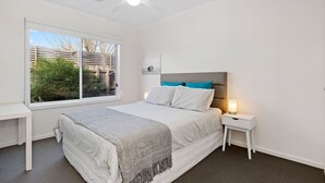 Un dormitorio con cama, mesita de noche y ventana con vista a árboles y una valla.