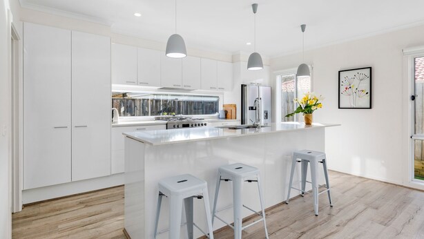 Una cocina moderna con armarios blancos, una isla central con taburetes, iluminación colgante y vistas al exterior.