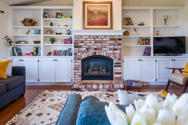 Uma sala de estar com lareira de tijolos, estantes brancas com livros e objetos decorativos, um sofá e uma televisão.