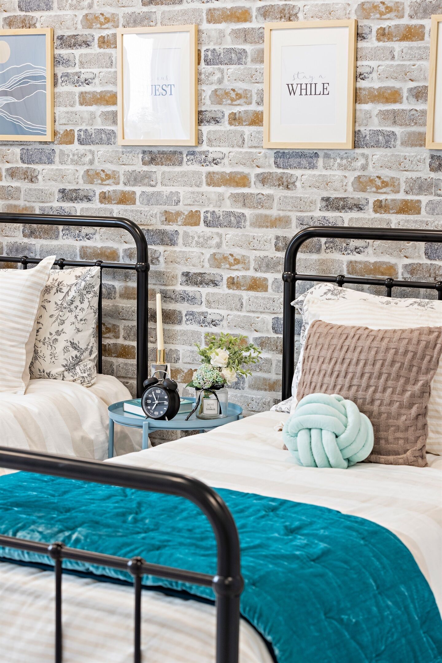A bedroom with two beds, a brick wall, framed artwork, a bedside table with a clock, and a decorative plant.